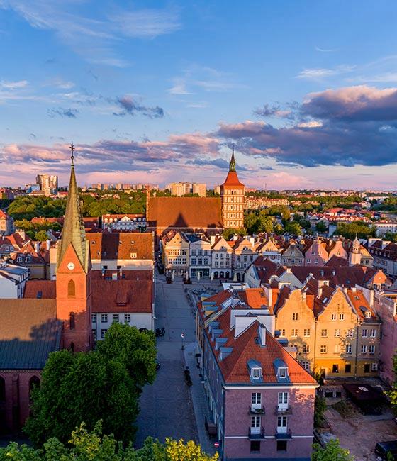 Panorama Starego Miasta w Olsztynie o zachodzie słońca – widoczne charakterystyczne zabytkowe kamienice, wieże kościołów oraz zieleń w tle.